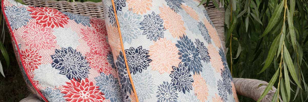 Close up image of two molly mahon cushions made from her dahlia fabric design with piped edges on a chair outside underneath a willow tree.