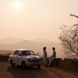 A vintage white car parked outdoors on a dirt road beside two people standing and sitting against a warm, hazy sunset sky with distant mountains and trees.