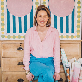 molly mahon sitting in her studio at home with her Bloomsbury painted pattern behind her and surrounded by her books, paint pots and wooden blocks