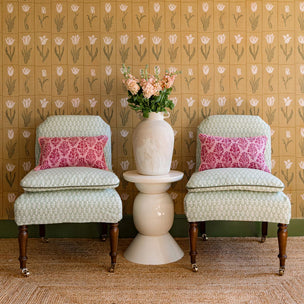 Two patterned chairs with pink cushions and a white vase on a small table against a floral wallpapered wall.