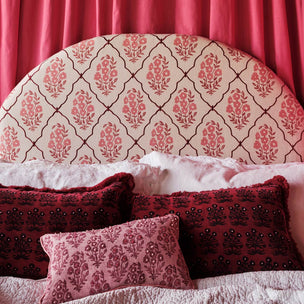 A close-up of a bed with a large upholstered headboard featuring a cream background and a pink damask-style floral trellis pattern. The bed is styled with deep red patterned pillows and a pink decorative pillow, set against a backdrop of rich red curtains.