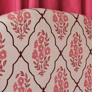 Close-up of upholstered fabric with a cream background, dark brown trellis pattern, and pink floral motifs, with a pink curtain visible at the top.