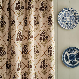 A close-up of a curtain panel with a cream background, brown floral motifs, and a tan trellis pattern, hanging next to two blue-and-white decorative plates on a light wall.