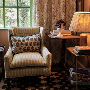 A classic upholstered armchair with cream and tan striped fabric and a matching patterned throw pillow, styled in a cozy room corner next to a wooden side table and a lit lamp.