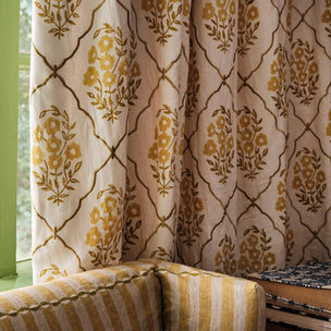 Patterned curtain panel hanging in a window, featuring a cream background with a mustard yellow floral trellis design. A matching striped armchair is partially visible in the foreground.