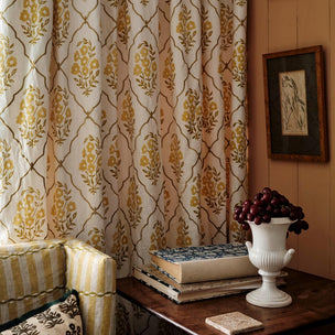 A styled interior scene showing patterned curtains in cream, tan, and mustard yellow hanging beside a wooden table with books, a white urn vase of dark grapes, and part of a striped armchair. A framed print hangs on the wall.