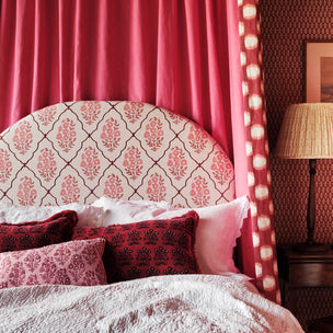 A styled bedroom scene featuring a bed with a cream upholstered headboard patterned with pink damask motifs, surrounded by pink and red patterned pillows, pink curtains, and a bedside lamp.