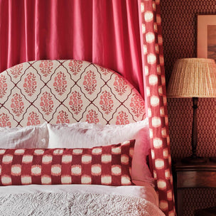 A styled bedroom scene featuring a bed with a patterned headboard and matching red-and-cream bedding, set against a red patterned wall with a lit table lamp on a bedside table.
