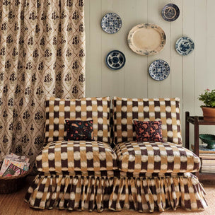 A two-seater sofa with a brown and beige checkered pattern, matching ruffled skirt, and two floral accent pillows, set in a styled room with patterned curtains, decorative plates on the wall, and a side table.