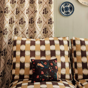 A styled interior scene showing patterned upholstery and curtains. The foreground is dominated by a sofa with thick back cushions in a vertical ikat-style pattern of brown, beige, and cream.