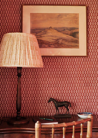 A framed landscape painting hanging on a wall with red patterned wallpaper, next to a table lamp and a small horse figurine.