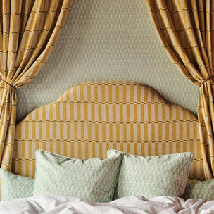 A styled bedroom scene featuring a bed with a curved upholstered headboard in a mustard yellow and cream striped fabric, flanked by matching curtains. The bed is dressed with two sage green patterned pillows and white bedding, set against a sage green patterned wallpaper.