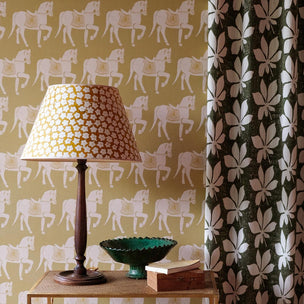 A styled interior corner showing patterned wallpaper with repeating horse motifs, a table lamp with a matching patterned shade, a dark green curtain, and a small side table with books and a bowl.