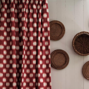 A rust red curtain panel with a white ikat-style square pattern hangs next to a white paneled wall decorated with three woven wicker baskets.