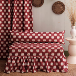A bed dressed in a rust red and cream ikat patterned bed skirt and matching pillowcase, styled in a rustic room with a matching curtain, a side table, and woven wall decor.