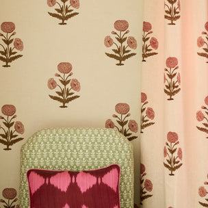 A styled interior scene showing floral wallpaper and a matching curtain in soft blush pink, with a green patterned chair and a pink accent pillow in the foreground.